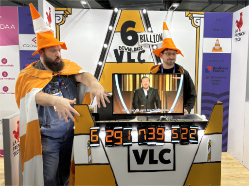 A photo of the VLC CES booth, with a big stilised orange traffic cone in the background, lights on the sides of the cone, text on it reading 6 BILLION DOWNLOADS VLC. In the foreground is a table with cones on the edges, a monitor on it displaying a video with subtitles autogenerated by VLC media player. Below the monitor, at the edge of the table are hanging several phones, each displaying a digit of a number, the count of VLC downloads, reading 6.291.739.522. On the left in front of the table is Julien standing, wearing an orange cone hat and a traffic-cone pattern cape. On the right, behind the table is jb standing, also wearing an orange traffic cone hat.