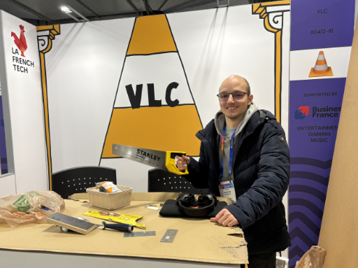 A photo of the VLC CES booth, with a big stilised orange traffic cone in the background, the booth visibily still unfinished with several tools on the table. Behind the table Marvin is standing, holding a saw in his right hand.