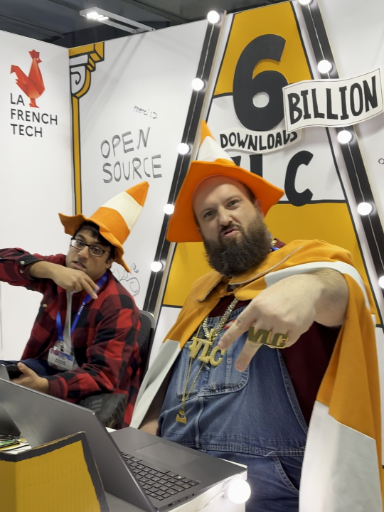 A photo of Kieran and Julien sitting at the VLC CES booth, both of them wearing orange traffic cone hats and Julien also wearing a traffic cone pattern cape.
