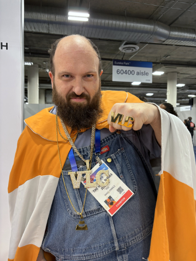 A photo of Julien wearing a traffic cone pattern cape, holding up his fist with big rings with golden letters on his fingers, forming the word VLC. He is also wearing a golden necklace with big VLC letters on it.